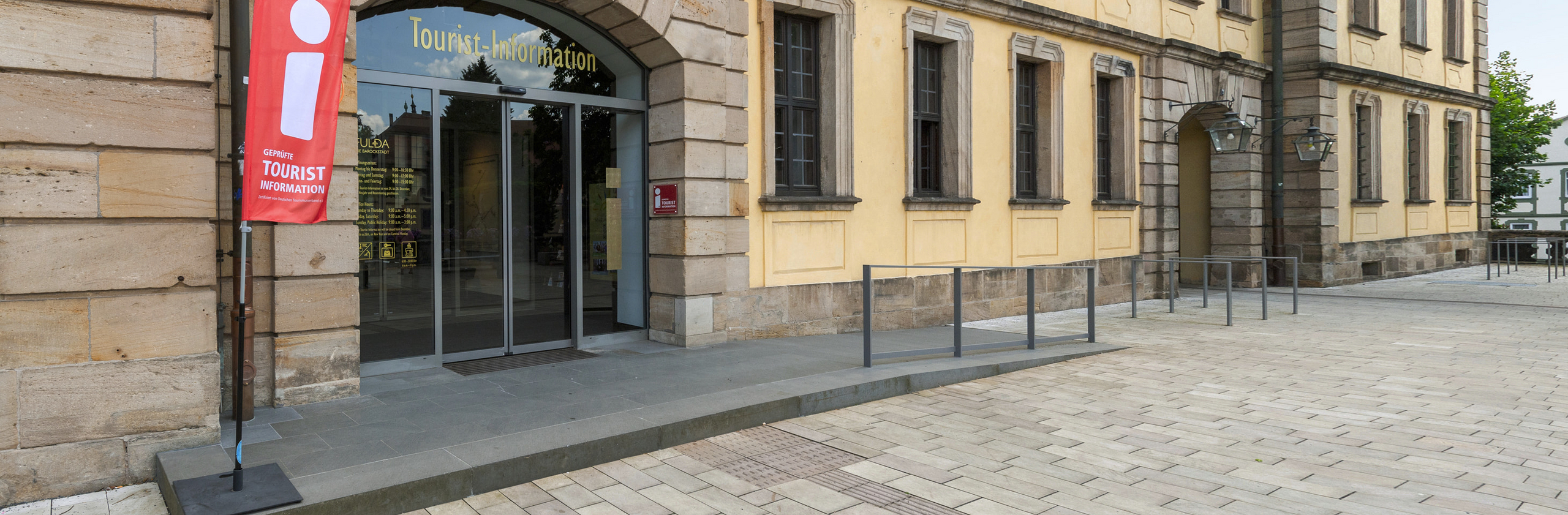 Eingangsbereich der Tourist-Information Fulda mit Rollstuhlaufgang und rotem Banner neben dem Eingang.